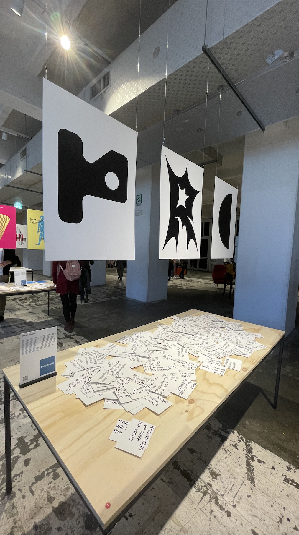 Installazione d'arte con forme geometriche sospese e tavolo con cartoncini scritti, in galleria espositiva moderna.