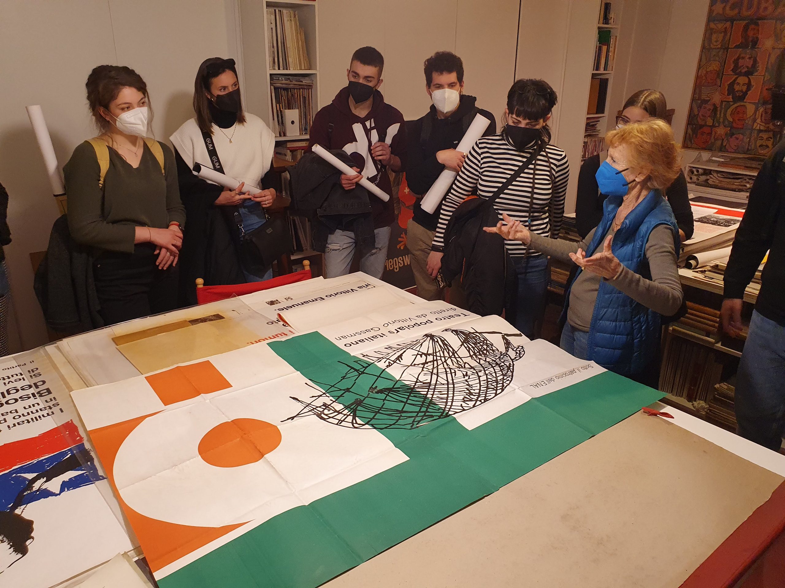 Gruppo di persone osserva e discute manifesti su tavolo in un laboratorio, alcune di loro indossano mascherine.