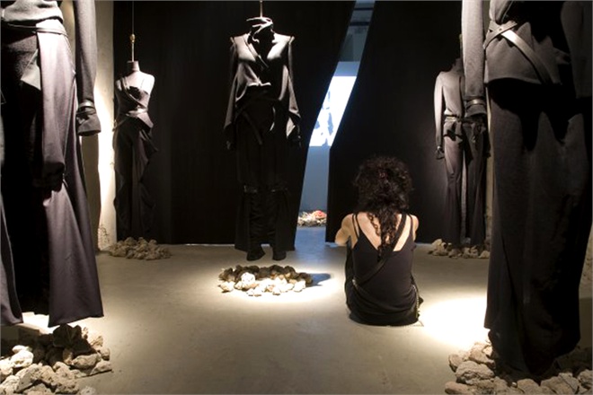 Mostra di moda con abiti neri sospesi e donna seduta osservandoli in una stanza illuminata.