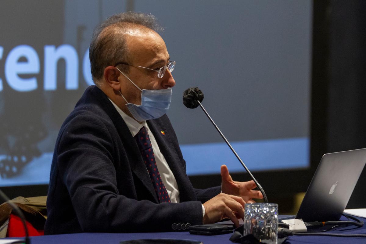 Uomo con mascherina parla al microfono durante una conferenza, laptop e bicchiere in primo piano.