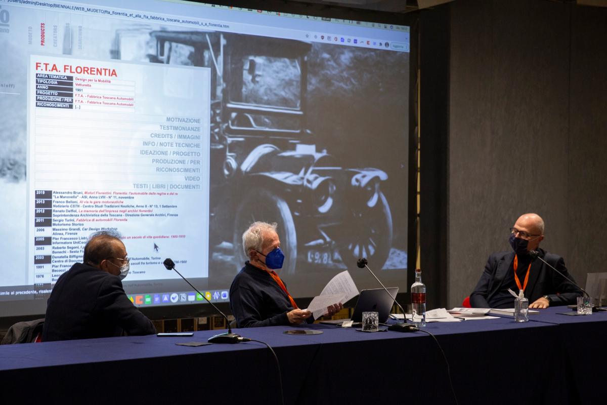 Tre uomini con mascherine discutono davanti a un grande schermo durante una conferenza sulla Fabbrica Toscana Automobili.