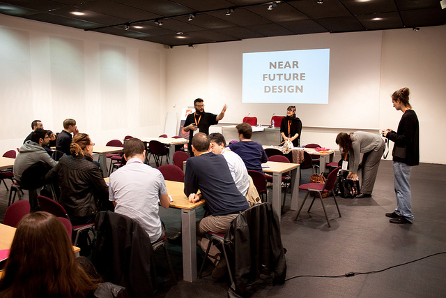 Una presentazione su Near Future Design in un'aula con persone sedute e un oratore in piedi.