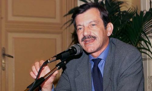 Uomo in giacca e camicia parla al microfono durante un evento al chiuso, sfondo con pianta e porta.