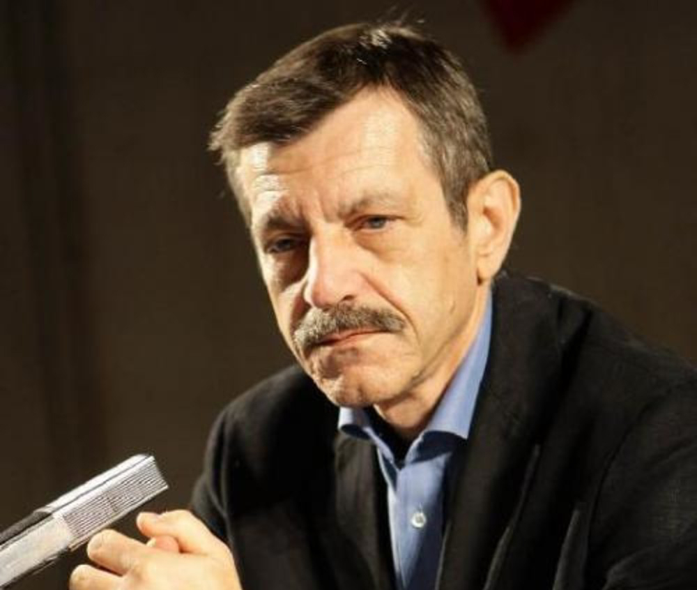 Uomo in giacca nera e camicia blu, seduto, tiene un microfono in mano durante un incontro pubblico.