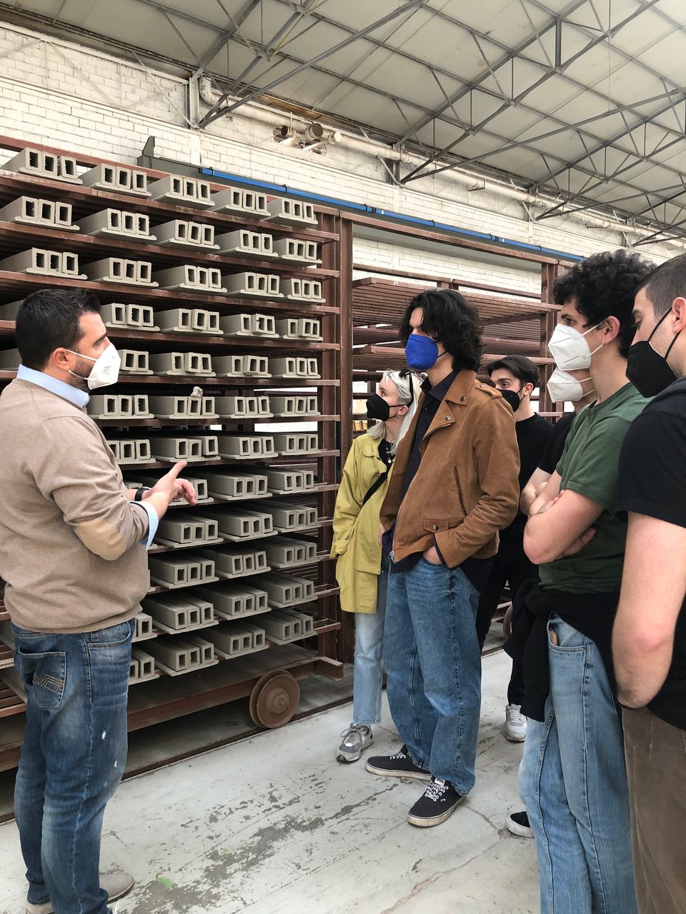 Gruppo di persone con mascherine visita una fabbrica di blocchi di cemento, ascoltando una spiegazione didattica.