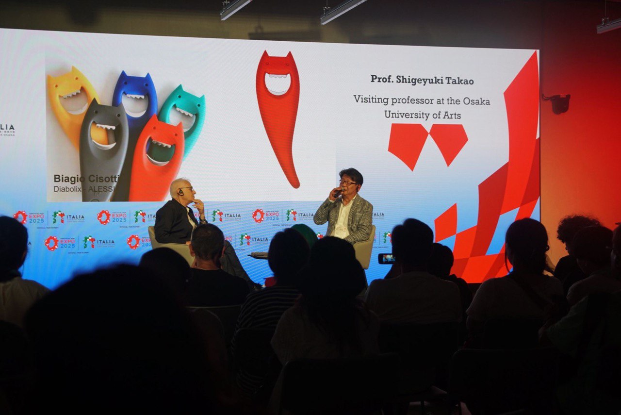 Due uomini seduti discutono in una conferenza con un pubblico, sfondo con grafica colorata e loghi Expo 2025.