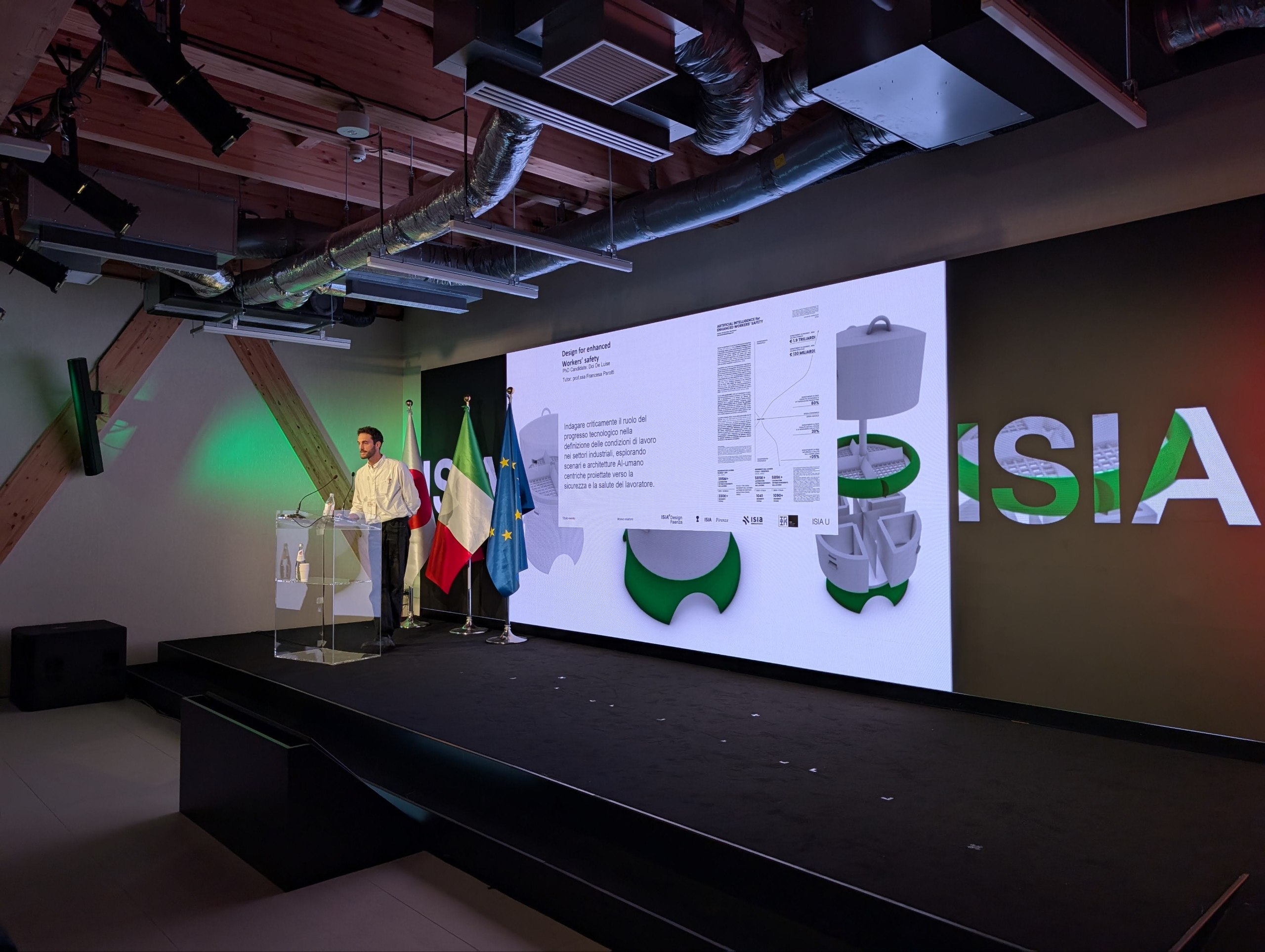 Oratore presenta su palco con schermo LED, bandiere internazionali e scenografia moderna industriale.