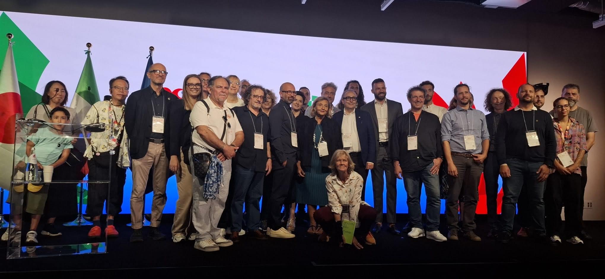 Gruppo di persone in posa davanti a bandiere italiane e giapponesi, sul palco durante un evento.