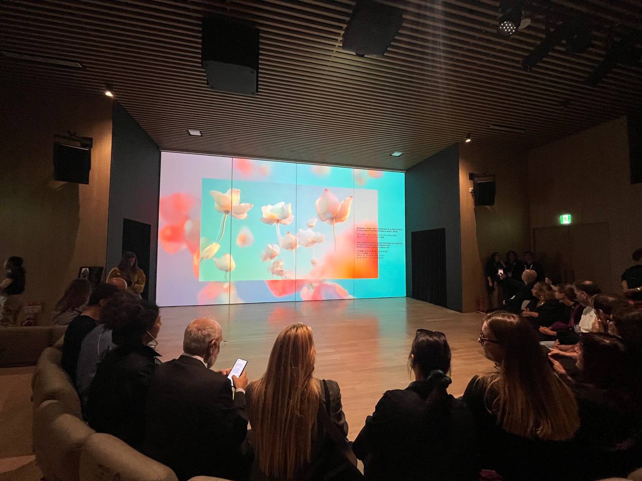 Persone sedute in una sala, osservano una grande proiezione luminosa di fiori su uno schermo, ambiente moderno.