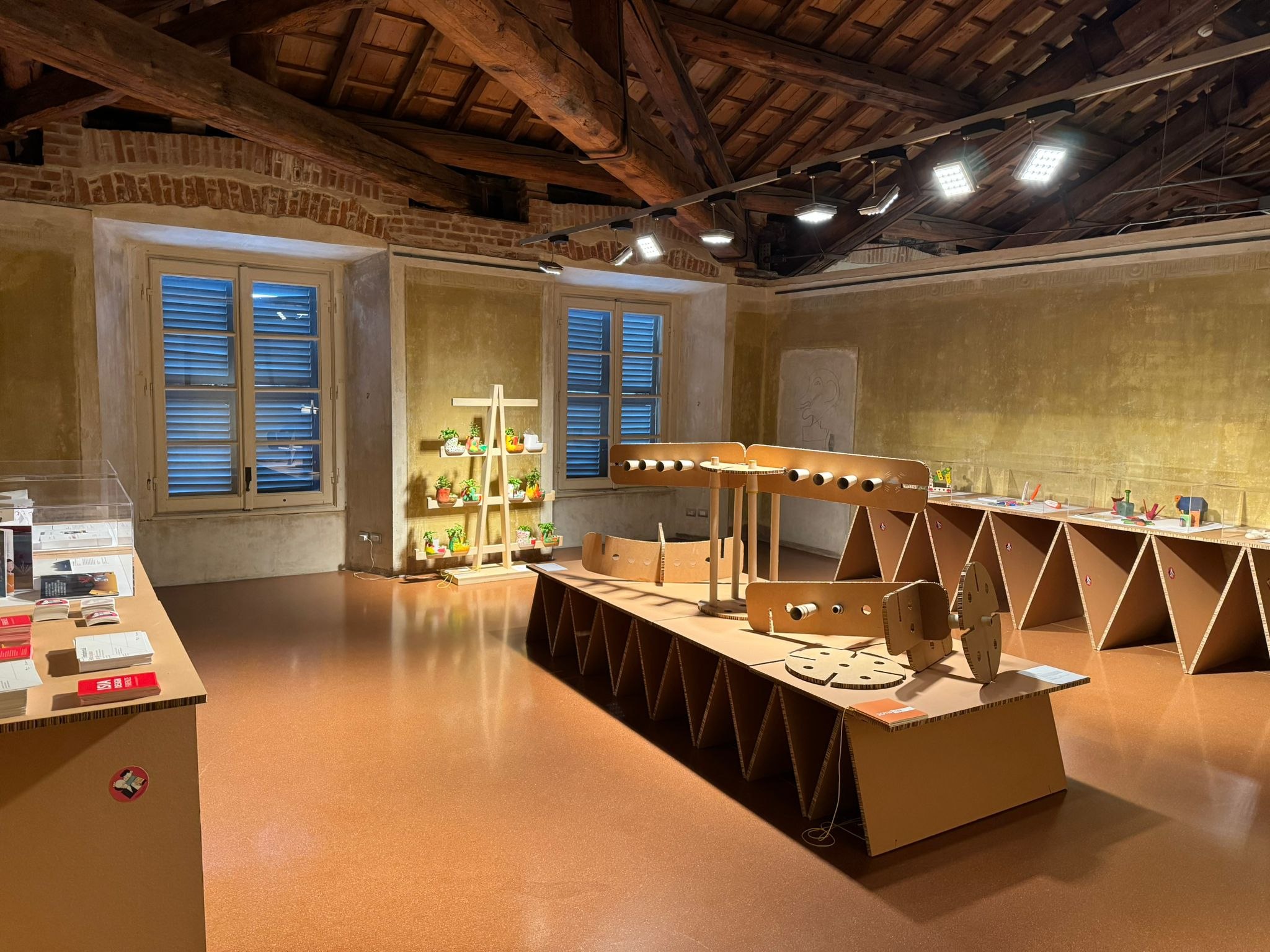 Interno di un museo con esposizione di oggetti in cartone e piante su scaffale, illuminato da luci soffuse.