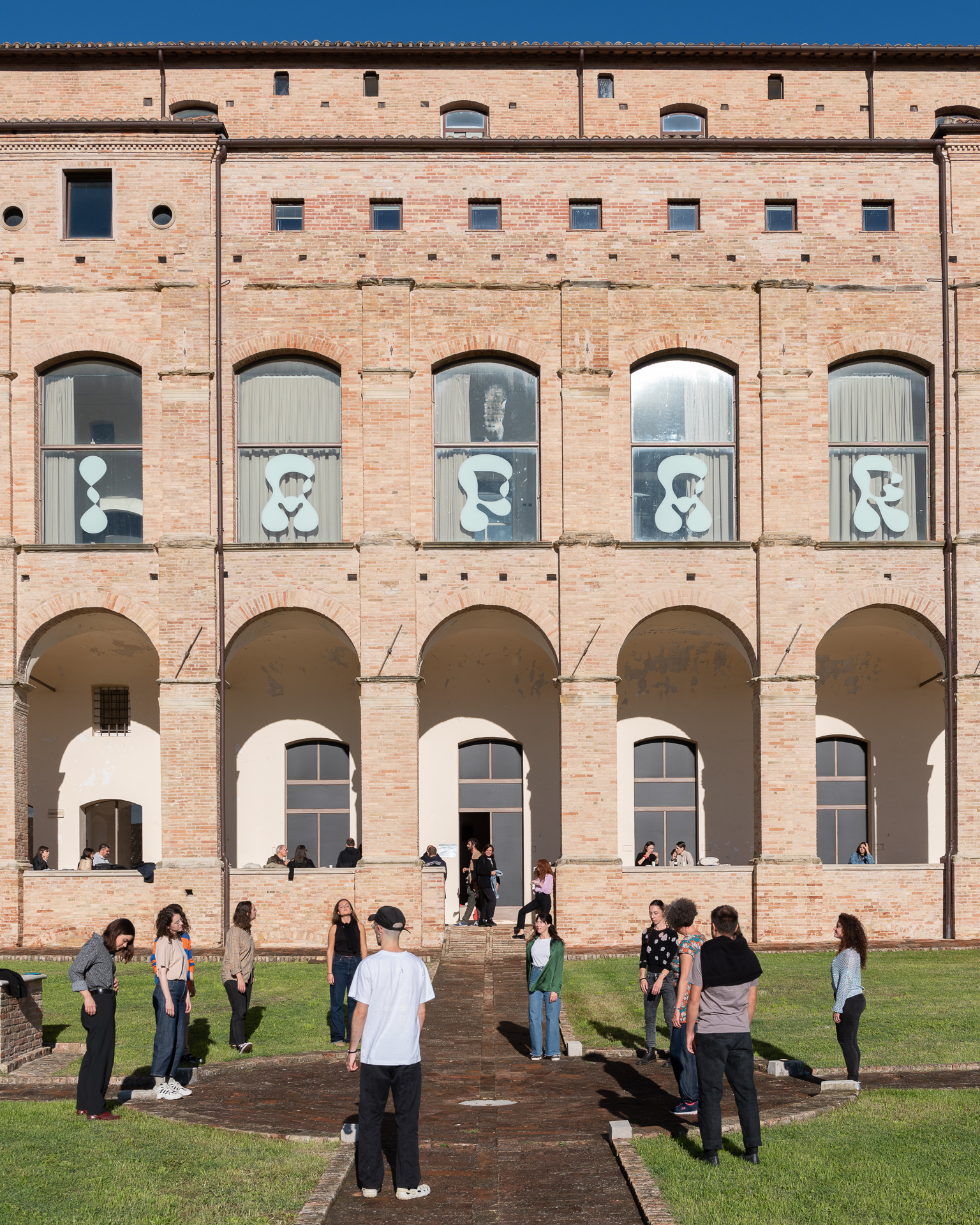 Gruppo di persone davanti a un edificio storico con grandi finestre e sculture grafiche, cielo azzurro.