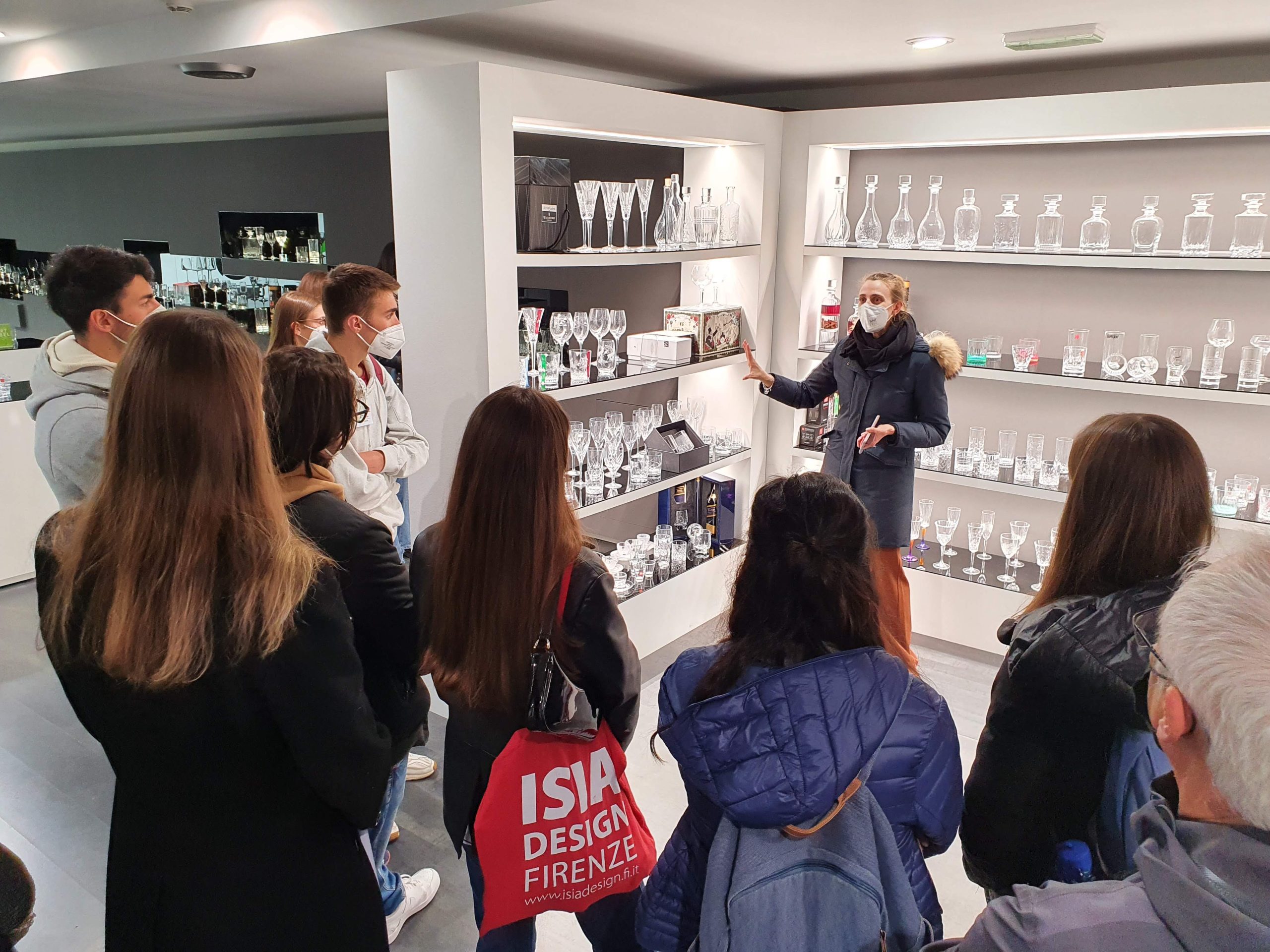 Gruppo di persone ascoltano una guida in un negozio di bicchieri di cristallo, indossando mascherine.