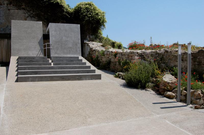 Scala di cemento moderna con pareti verticali, cielo azzurro e giardino fiorito lungo un muretto di pietra.