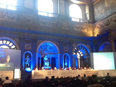 Conferenza UNESCO in una sala storica con relatori e pubblico, decorazioni artistiche sullo sfondo.
