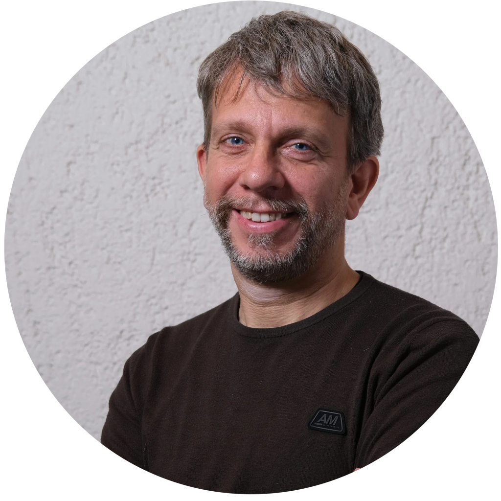 Uomo sorridente con capelli grigi e barba, indossa una maglia scura davanti a un muro bianco.