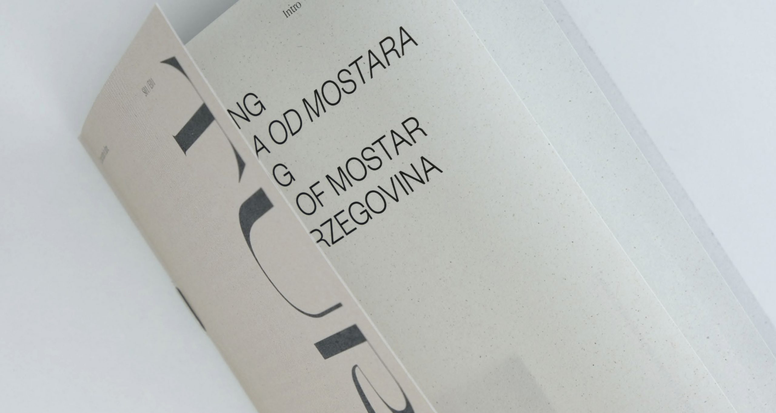 Pagina di libro con testo Mostar visibile, sfogliata da una mano su sfondo bianco minimalista.