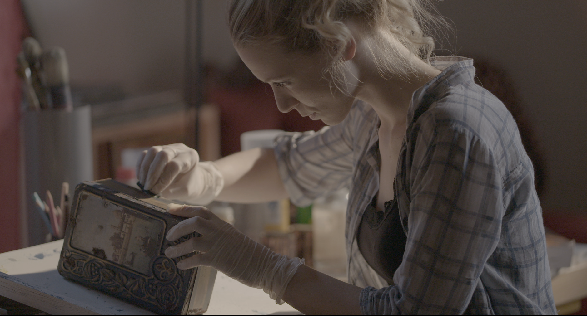 Donna concentrata restaura un'antica scatola, indossando guanti e camicia a quadri, in uno studio d'arte.