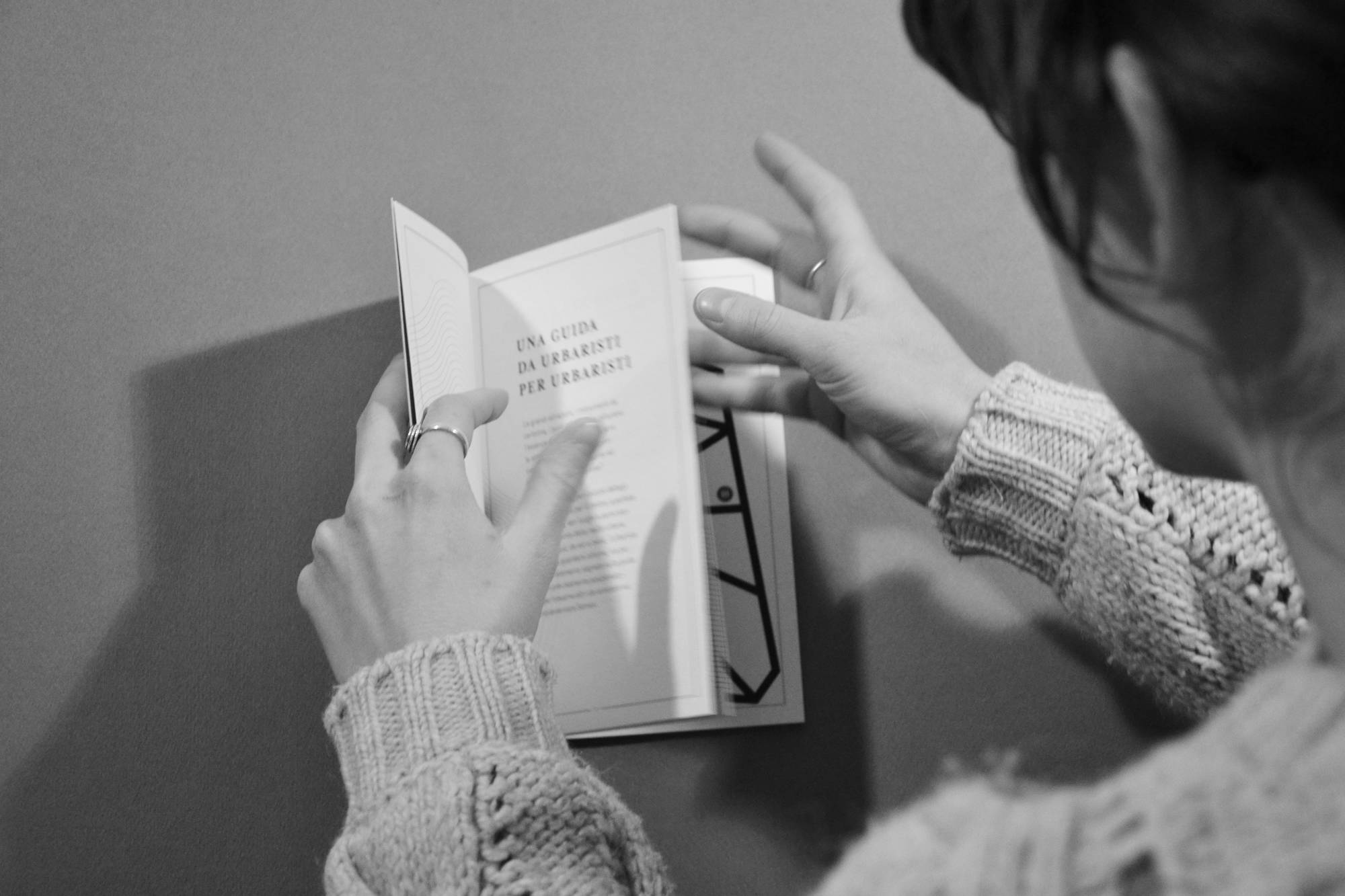 Persona sfoglia un libro dal titolo Una guida da urbanisti per urbanisti, indossando un maglione lavorato a maglia.