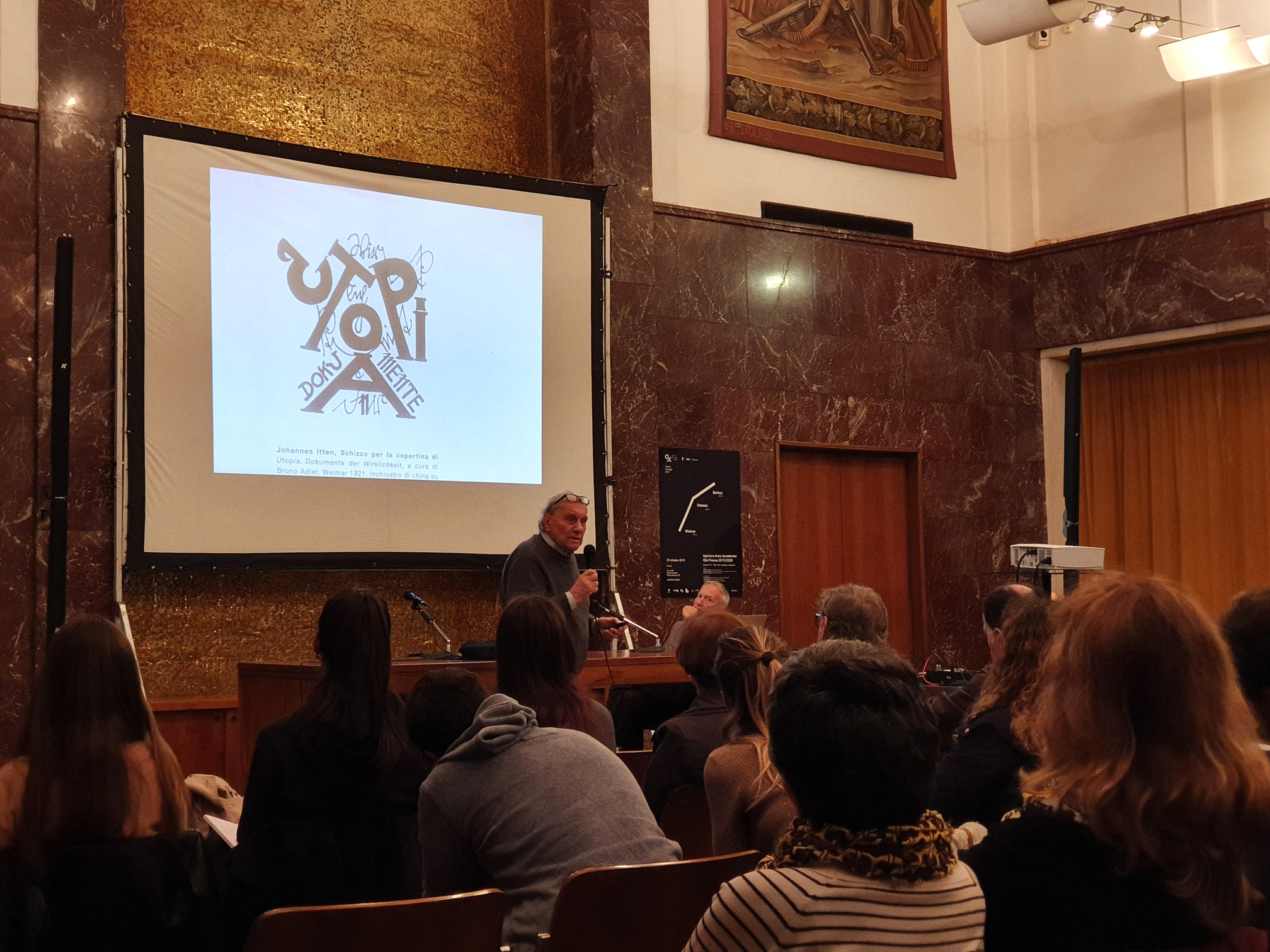 Conferenza in una sala con spettatori, relatore al microfono e schermo proiettato con un'opera grafica.