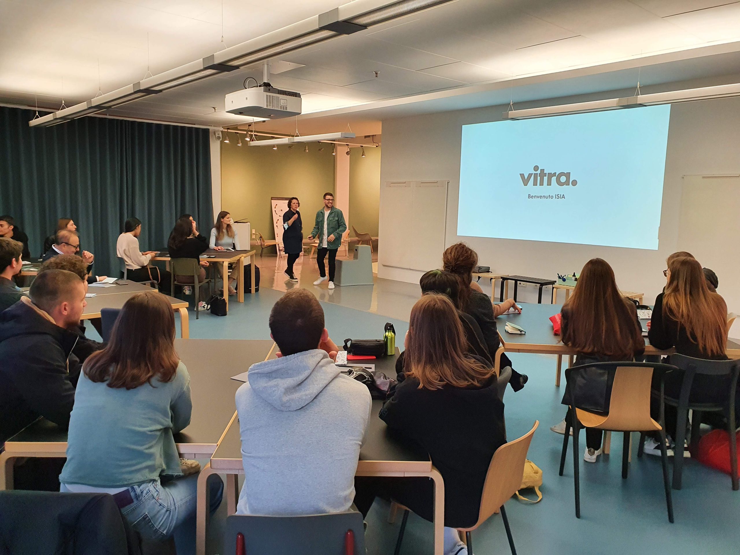 Persone in una sala conferenze partecipano a una presentazione con proiezione del logo vitra sullo schermo.