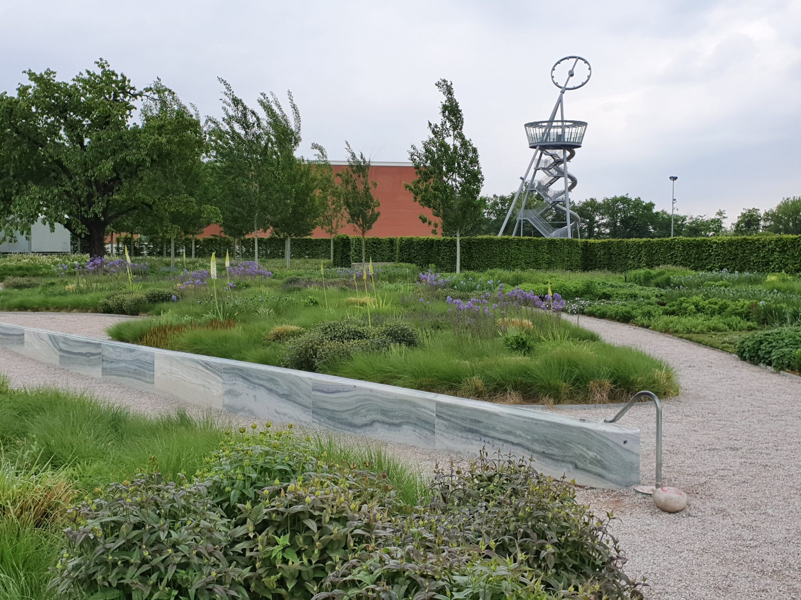 Giardino paesaggistico con sentiero curvo, piante verdi e scultura di metallo sullo sfondo.