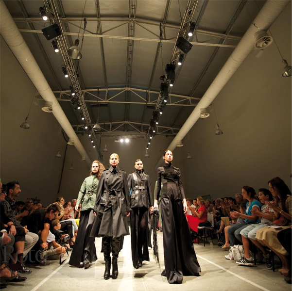 Sfilata di moda con modelli in abiti eleganti su passerella, pubblico assorto ai lati dell'evento.