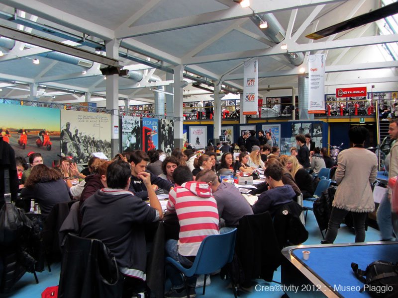 Gruppo di persone partecipa a un evento collaborativo al Museo Piaggio, circondato da poster e immagini storiche.